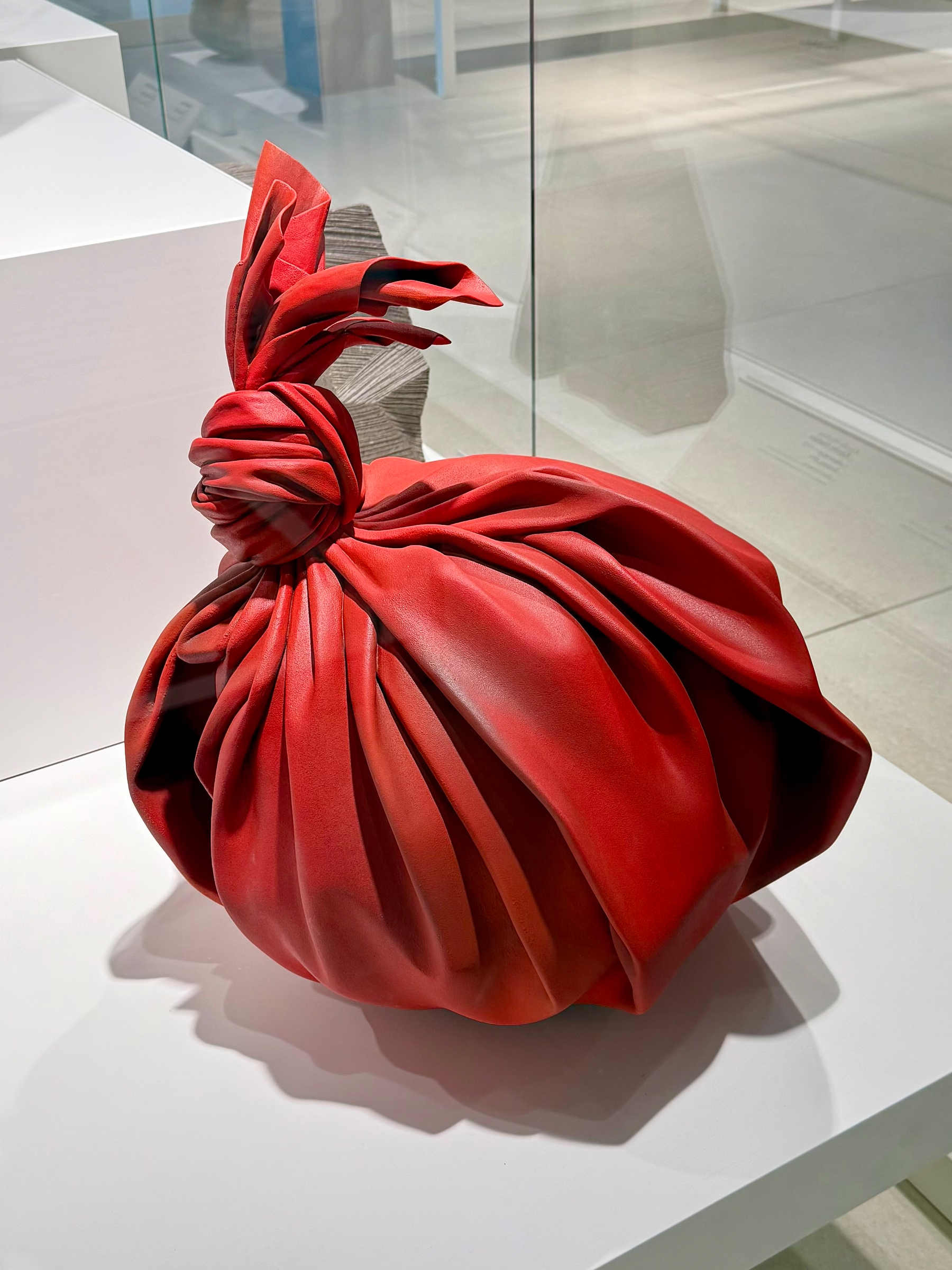 A sculpture resembling a large, intricately folded red bag with a tied top, displayed on a white surface in a minimalist setting. The material appears smooth and the shadows cast create a sense of depth and texture.