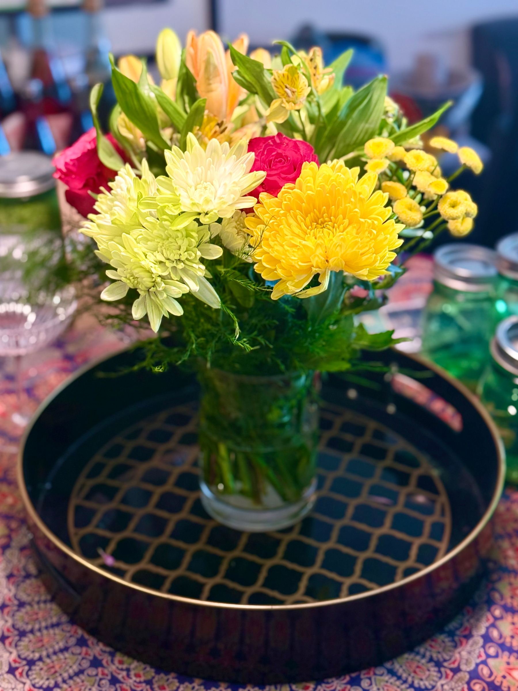 A clear glass vase holds a vibrant bouquet of mixed flowers, including bright yellow chrysanthemums, soft green chrysanthemums, pink roses, pale orange lilies, and small yellow button flowers, surrounded by lush green foliage. The vase sits on a round black tray with a gold geometric lattice pattern in the centre. The tray rests on a table covered with a colourful, intricate, floral-patterned tablecloth in shades of purple, pink, and cream. In the background, slightly out of focus, are glass jars, one green and one clear, along with other household items. The scene is warmly lit, highlighting the vivid colours of the flowers.