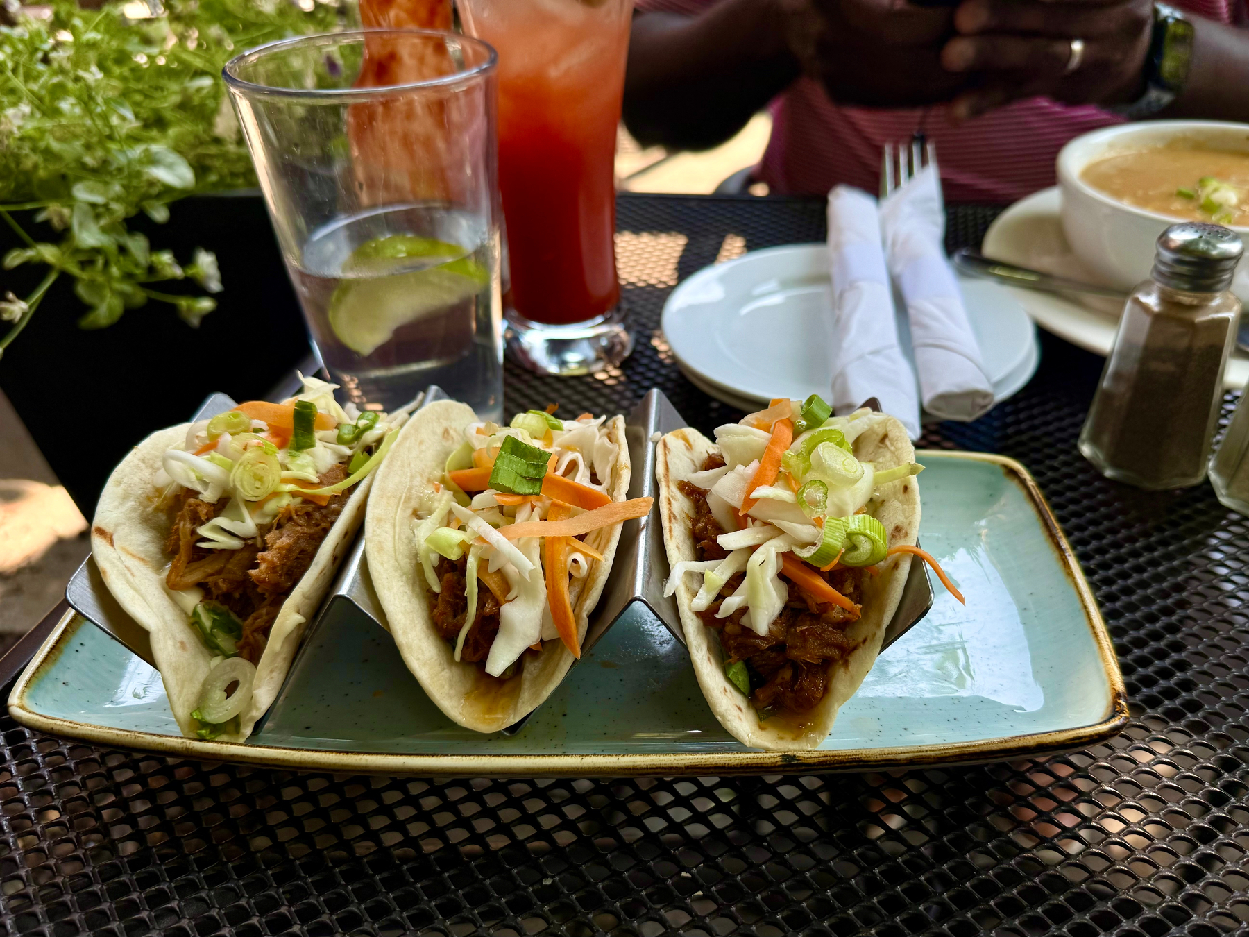 A plate with three soft tacos filled with shredded meat, shredded cabbage, carrot strips, and chopped green onions sits on a black metal patio table. The tacos are held upright in a metal taco holder on a rectangular light blue plate with a brown edge. Behind the tacos are a glass of water with a lime wedge, a tall red drink with ice, and a bowl of soup on a white saucer with a spoon and a pair of rolled white napkins on a separate white plate. A pepper shaker is nearby, and a person wearing a red and white striped shirt is holding a phone in the background. Green plants are visible in the top left corner.