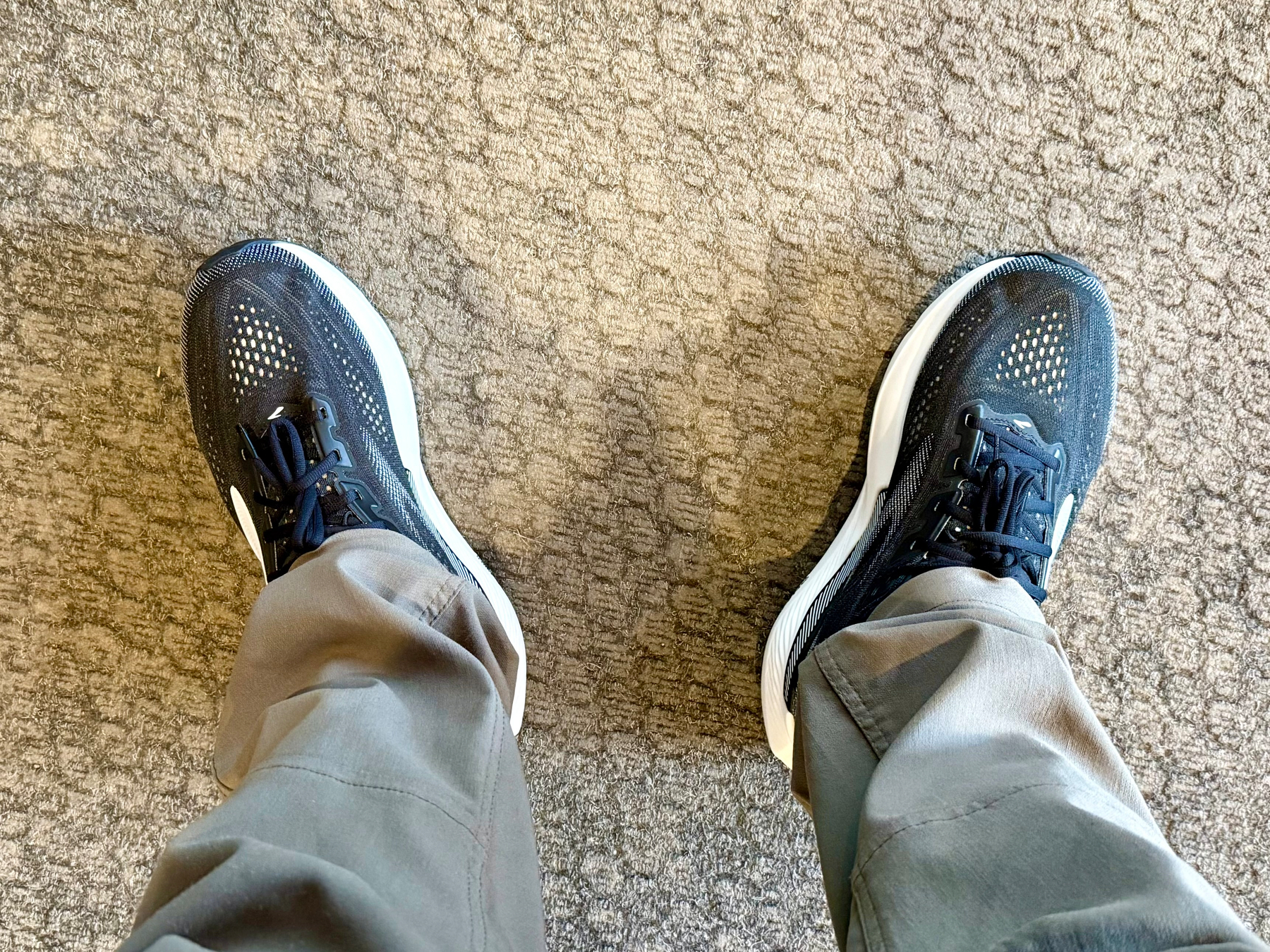 A person is standing on a patterned beige carpet, wearing light grey pants and black athletic shoes with white soles. The shoes have a mesh upper with small perforations and black laces. The photo is taken from the person’s perspective, looking down at their feet, with both legs slightly apart.