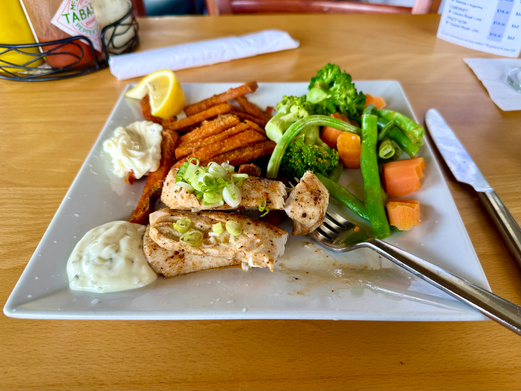 A square white plate holds a meal consisting of grilled fish fillets topped with chopped green onions, sweet potato fries, and a mix of steamed vegetables including broccoli, green beans, and carrot chunks. Two dollops of white sauce are on the plate, along with a lemon wedge. A fork and knife rest on the right side of the plate. In the background, there are condiment bottles in a wire holder, including a bottle of Tabasco sauce, and a menu is partially visible on the wooden table.