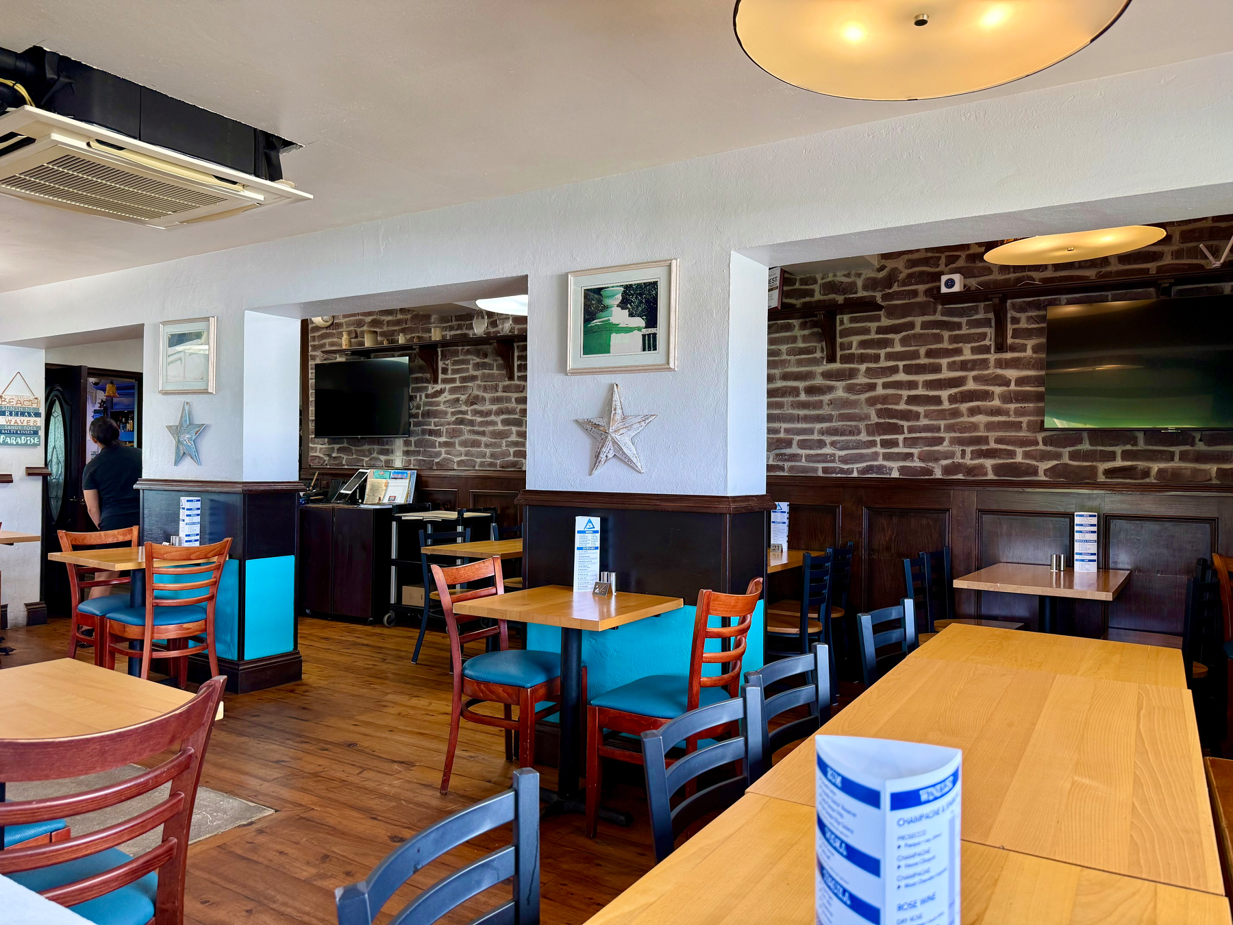A cozy restaurant interior featuring wooden floors and tables with a mix of wooden chairs, some with teal cushions and others with black or dark wood finishes. The walls are partly white and partly exposed brown brick, with dark wood paneling along the lower sections. A few framed pictures and metallic star-shaped wall decorations hang on the white walls. Two flat-screen TVs are mounted on the brick walls in the back sections of the dining area. Ceiling lights with round, flat fixtures provide warm illumination, and a visible air conditioning unit is mounted on the ceiling. A staff member dressed in black is seen walking toward a door on the left side of the image. Several tables display folded blue-and-white paper menus.