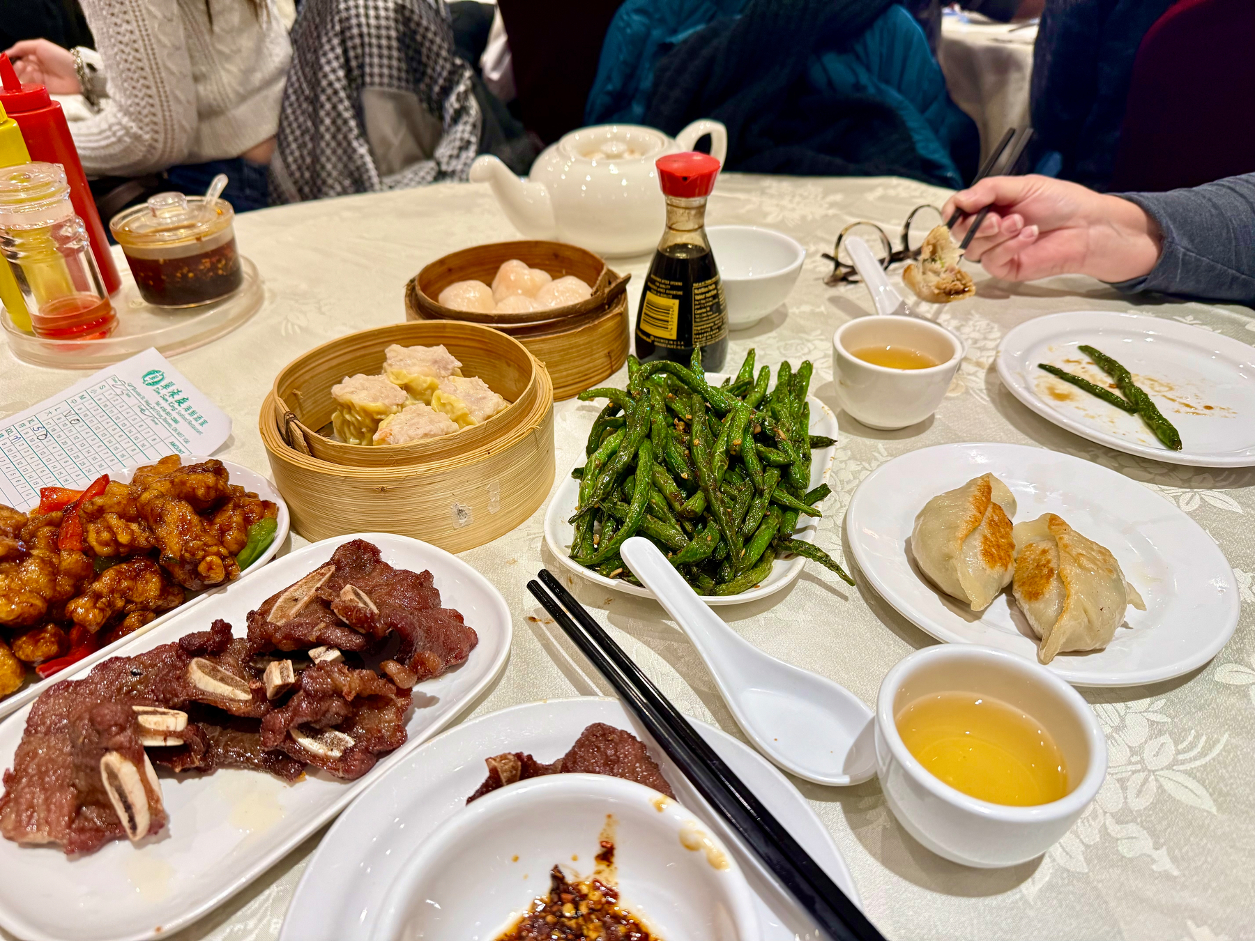 A table set with a variety of Chinese dishes, including a plate of grilled beef ribs, sweet and sour chicken, stir-fried green beans, and two steamed buns on a white plate. A bamboo steamer contains dumplings, and there is a soy sauce bottle nearby. Two cups of tea are on the table, along with a soy sauce dish, chopsticks, and a menu slip. A person is picking up a dumpling with chopsticks. A white teapot is also visible, and condiments like chili oil and sauces are in the background.