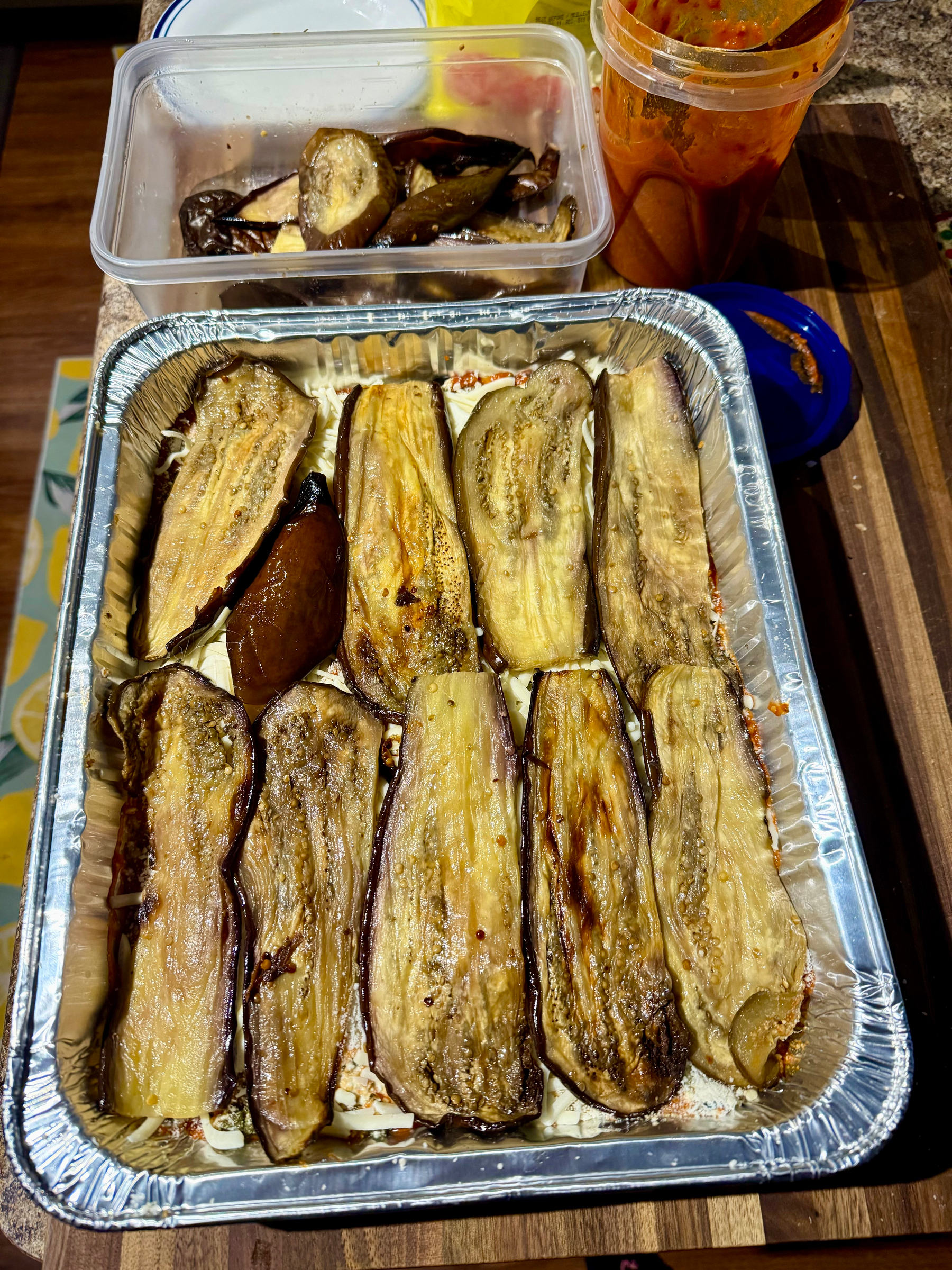 Roasted eggplant slices are layered in a foil tray, with a plastic container and a sauce container nearby.