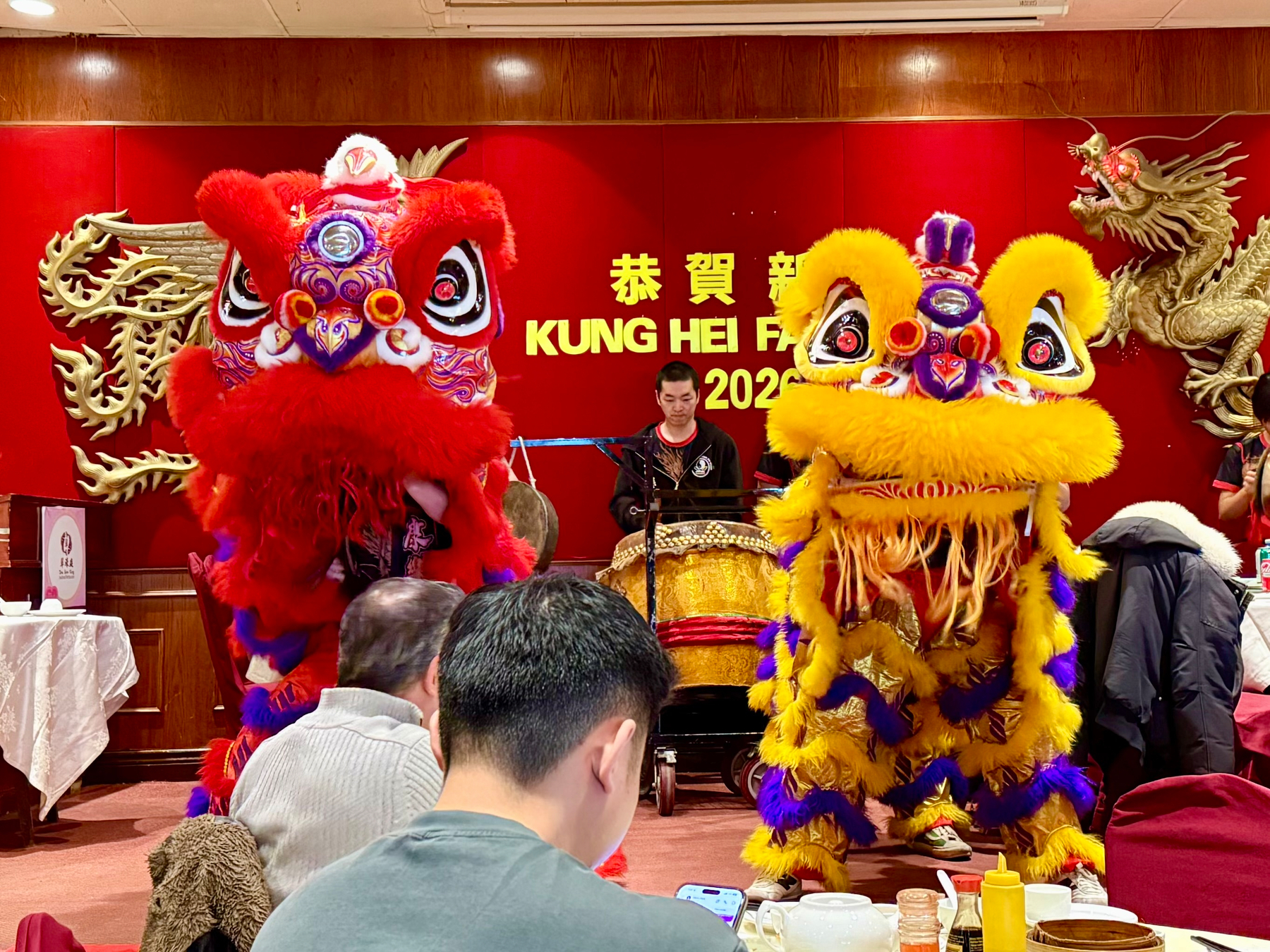 Lion dancers in vibrant red and yellow costumes perform at a festive celebration. One dancer in a red lion costume and another in a yellow and blue lion costume move energetically in front of a drummer. The background features red walls with golden Chinese dragons and a sign that reads "Kung Hei Fat Choi 2026." Several people are sitting at tables in the foreground, watching the performance.