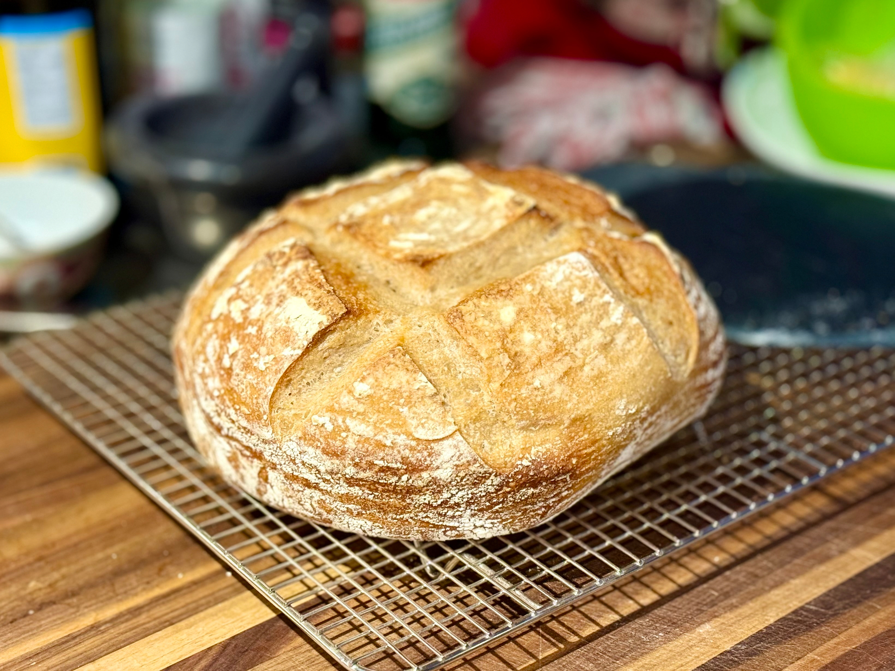 A freshly baked round loaf of artisanal bread rests on a wire cooling rack atop a wooden countertop. The bread has a golden-brown crust with a rustic, lightly floured surface and a distinctive scored pattern forming a large X across the top. The background is slightly blurred, showing kitchen items including a green mixing bowl, a white plate, a mortar and pestle, and other assorted containers. The bread appears crisp and airy with a well-developed crust.