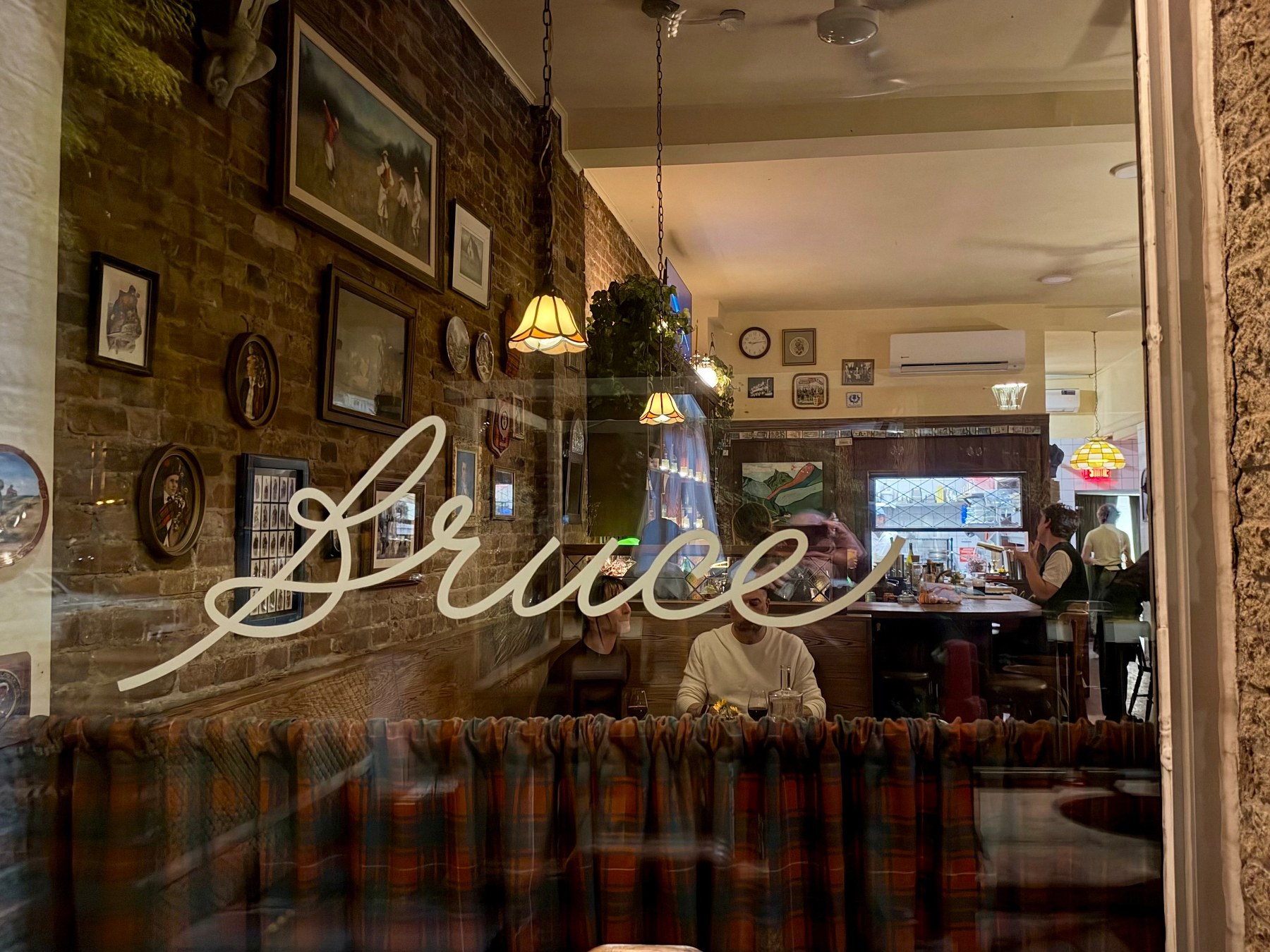 A window view into a warmly lit café with a brick wall on the left covered in framed pictures of various sizes. Hanging lamps with yellow shades illuminate the space. A person in a light top sits behind a plaid-patterned curtain along the window. In the background, a counter with a person standing and a sign reading “Bruce’s” are visible. The word “Bruce” is written in large white cursive across the glass.